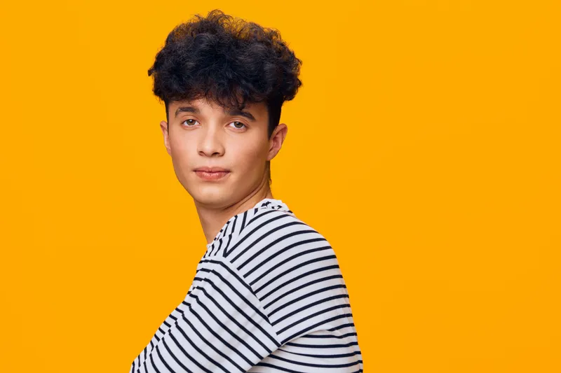 Young man with curly hair wearing striped shirt looks confidently at camera against vibrant orange background, casual style and soft lighting creating warm mood.