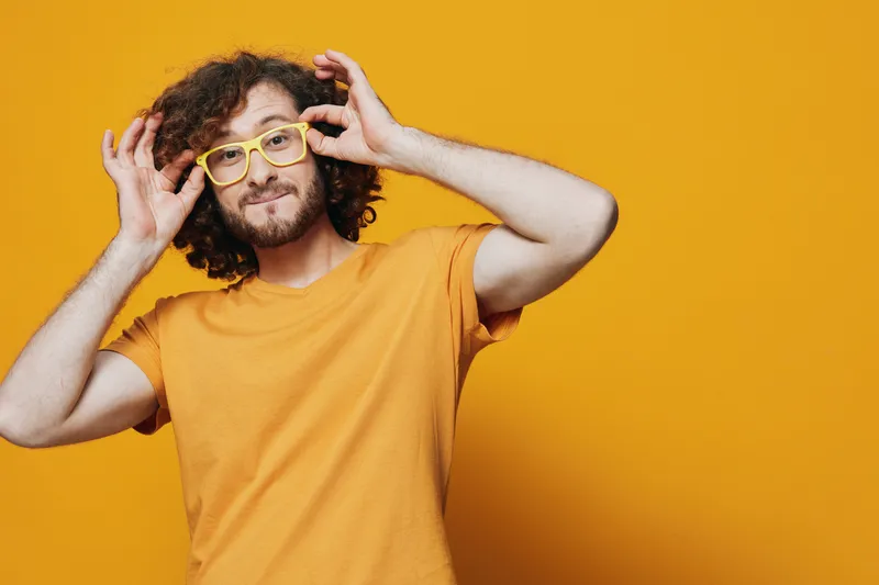 Cheerful young man with curly hair and glasses against a bright yellow background, expressing joy and positivity in a casual orange t shirt