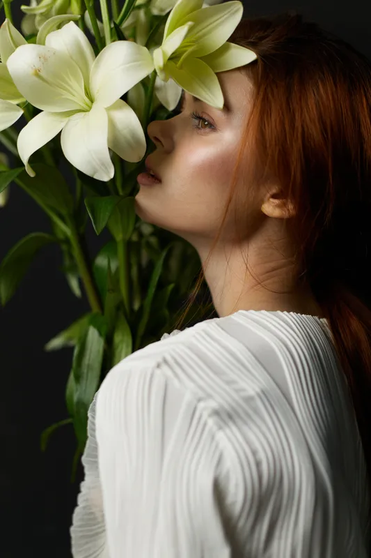 Young woman with red hair posing with white lilies against a dark background, showcasing elegance and tranquility, perfect for wellness and beauty concepts