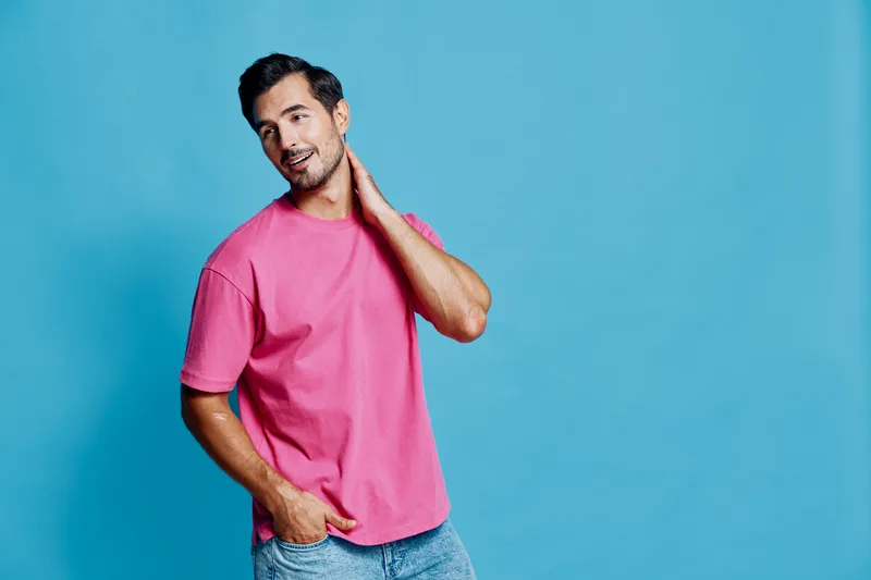 Smiling young man wearing pink t-shirt poses casually while looking at the camera, showcasing relaxed confidence against a vibrant blue background.