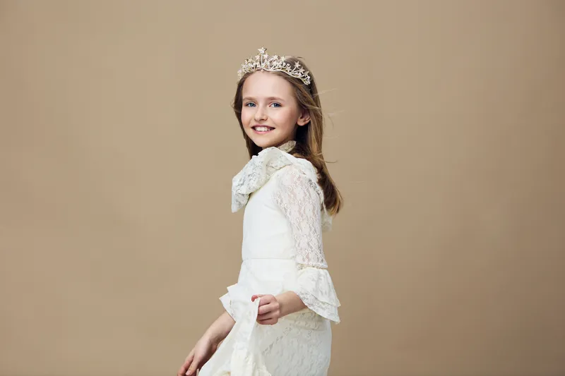 A joyful young girl wearing a white lace dress and a tiara, posing elegantly against a soft beige background, embodying grace and childhood wonder