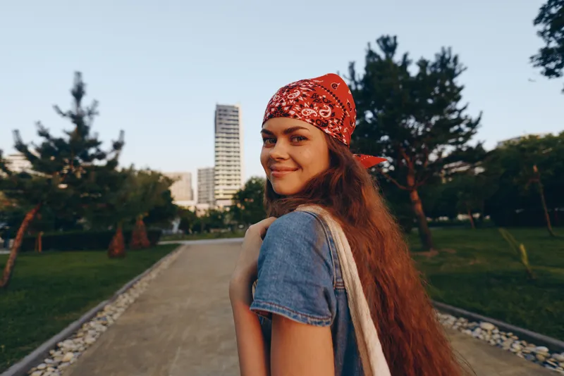 lifestyle woman in modern boho-western denim street style with red bandana, long hair, smiling outdoors in natural park setting under warm film color tones