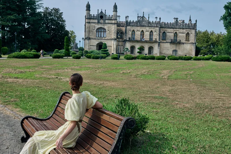A woman in a pale dress sits on a wooden bench beside a green lawn, admiring a grand historic castle and its manicured garden under a clear sky