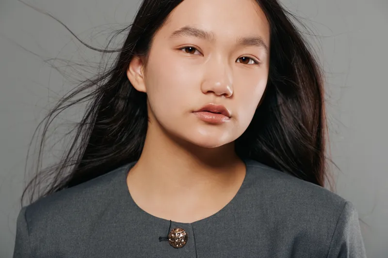 Woman with long dark hair and natural makeup wearing gray jacket and looking at camera on gray background. Studio portrait of confident female with calm expression and minimal styling.