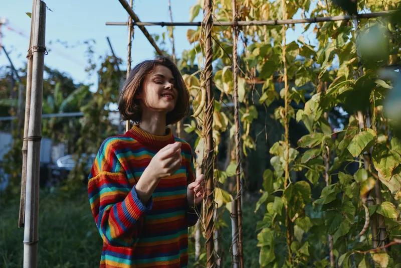 Woman in a colorful striped sweater smiles while touching climbing vines on a wooden trellis in a sunlit garden. Young adult with short hair in casual outfit, cozy lifestyle scene.