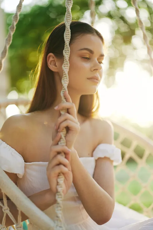 Young woman in casual white dress, conveying tranquility while resting on a swing, bathed in warm sunlight against a lush green background