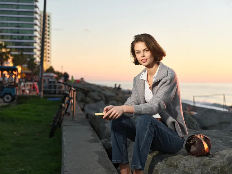 A thoughtful woman sits on a rocky shore at sunset, wearing a blazer and jeans, gazing toward the water with a calm, confident expression.