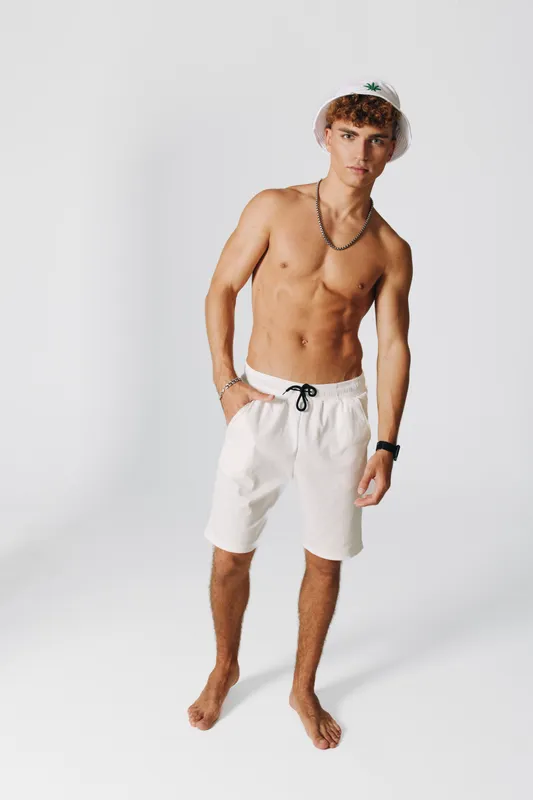 Young man standing bare chest with white shorts comfortable casual style accessories in living room with white background and relaxed confident posture full length