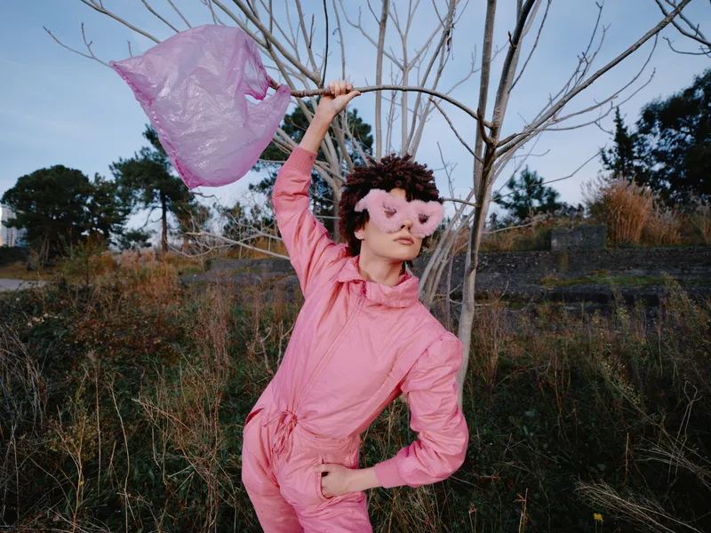Child girl in pink costume and mask dances outdoors with flowing fabric, capturing playful whimsy and imaginative performance in a natural field under soft light