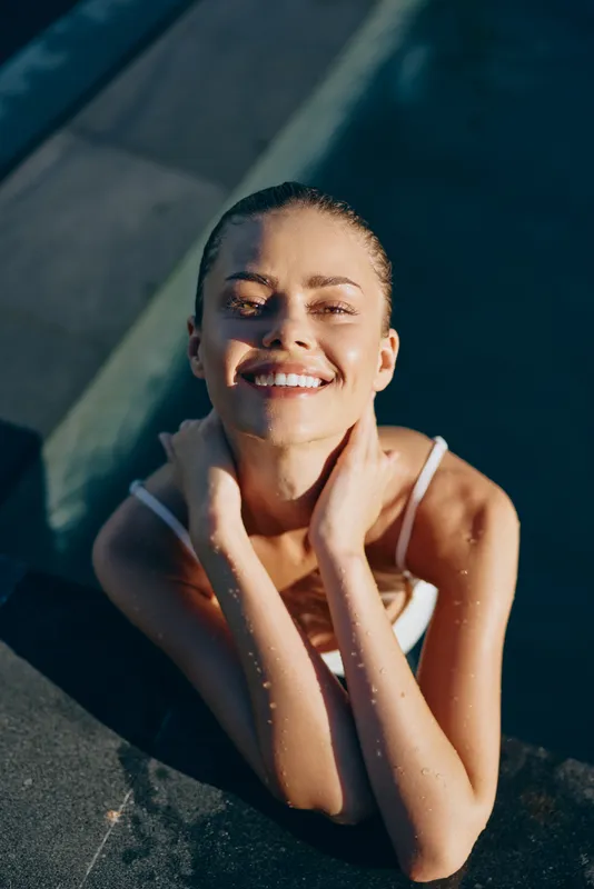 Smiling woman by a pool, leaning on the edge with hands at her neck, sunlight on skin, wet hair, warm mood, outdoor lifestyle, poolside relaxation, summertime wellness, carefree moment.