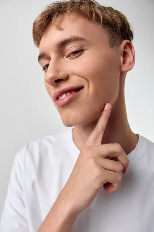 Young man thinking smile finger on chin casual white shirt confident relaxed studio lighting pensive expression thoughtful pose modern clean backdrop image