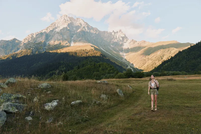 Woman hiker in a mountain meadow, exploring rugged terrain with a backpack, shorts and boots, gazing at distant peaks, a calm outdoor scene with solo travel vibe.