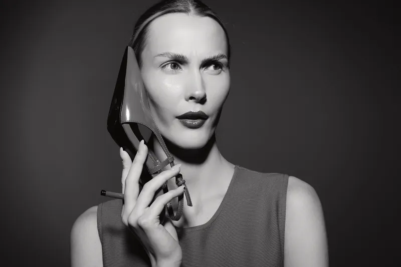 Portrait of a fair skinned Caucasian woman holding a high heel shoe close to her face, looking thoughtfully with sleek hair and bold lips in black and white.