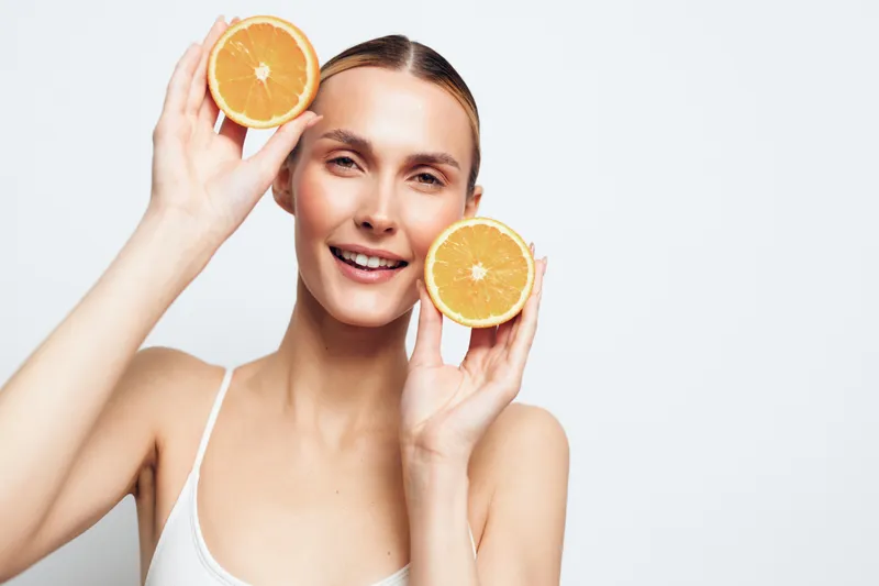 A smiling young woman holds orange halves near her cheeks in a bright studio, presenting a fresh, healthy lifestyle concept.