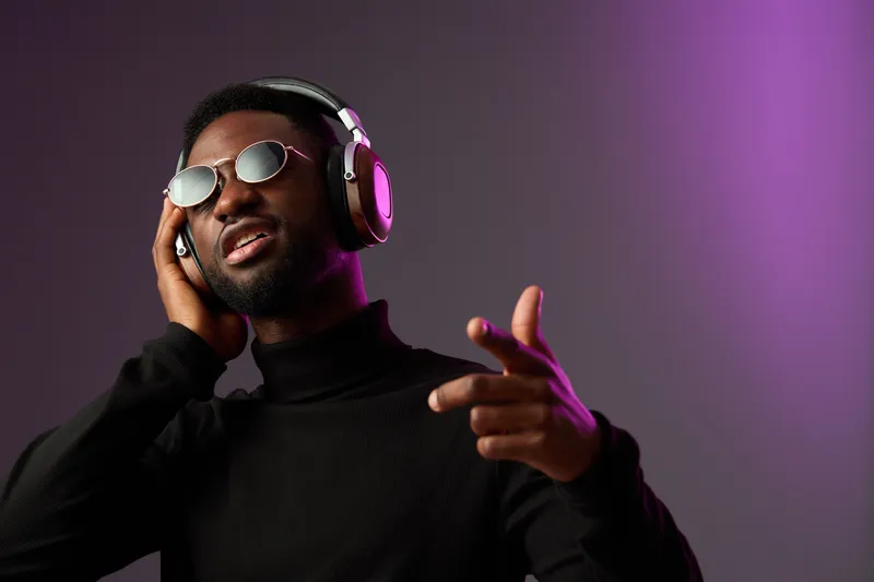 A stylish Black man in sunglasses enjoying music with headphones, expressing joy in a modern minimalist setting with purple lighting, perfect for music themes