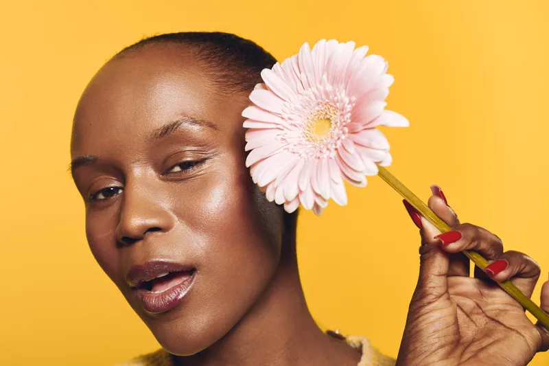 Close-up of a dark skinned African woman holding a pink flower near her face on yellow background. Concept of natural beauty, skincare, floral, wellness, and self-care with bright lighting.