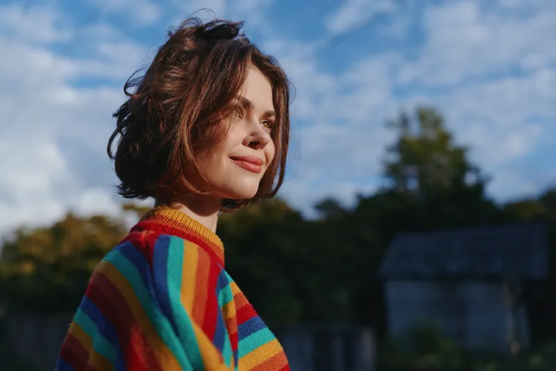 Woman, striped sweater, short hair, smiling outdoor portrait of a relaxed young woman in her twenties wearing casual rainbow knit, looking sideways in soft natural light for lifestyle.