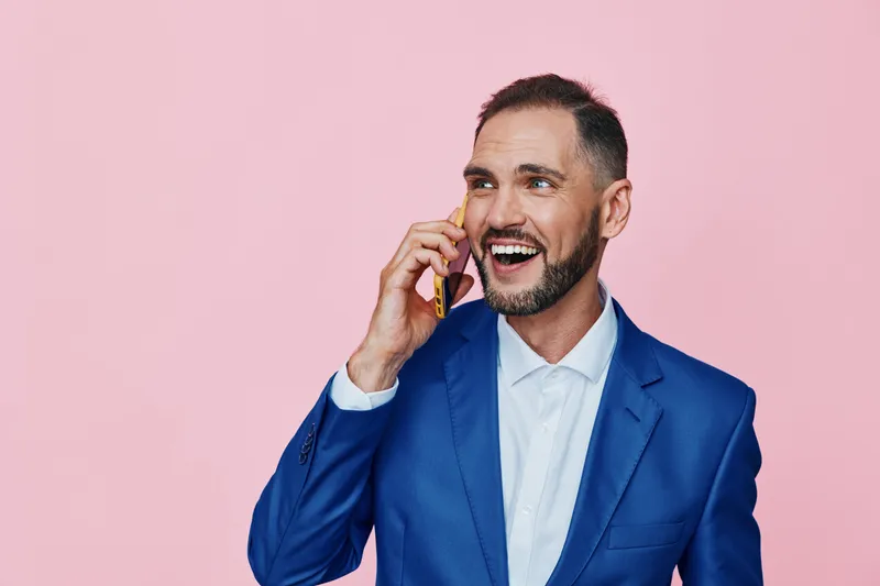A business professional man stands against a solid pink backdrop, engaging in a phone call with a confident smile, radiating positive energy and focused intent.