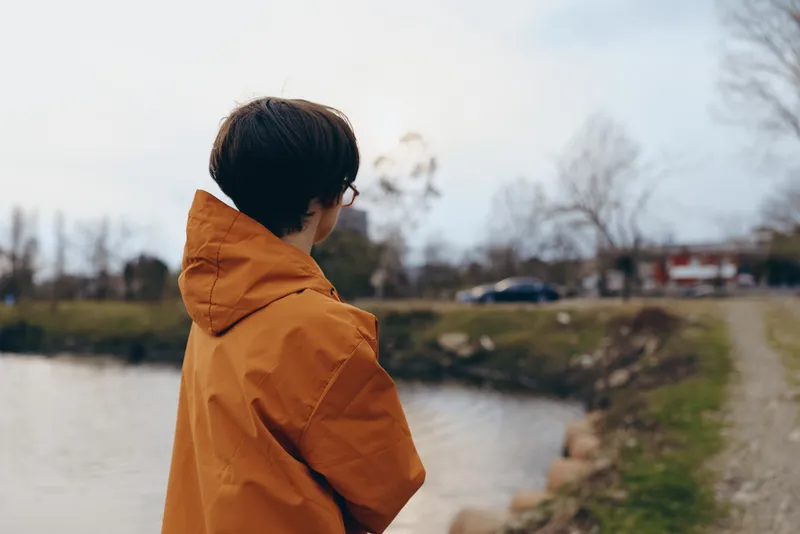 Woman lifestyle outdoors by river in winter, wearing orange hooded coat and hood, walking on riverside path, back turned with hands in pockets, calm peaceful mood, soft daylight with blurred trees,