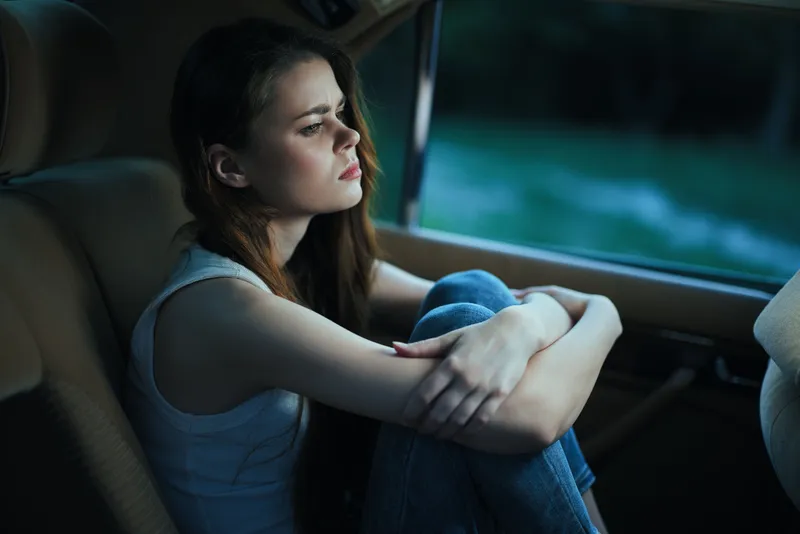 Pensive young woman in a car with a contemplative expression, dressed casually in a white tank top and jeans, reflecting on life.