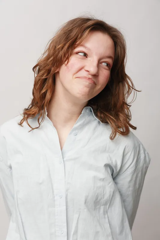 Young woman with curly hair wearing white shirt looks sideways with playful smile on light background. Concept of casual style, positive emotion, and natural beauty portrait.