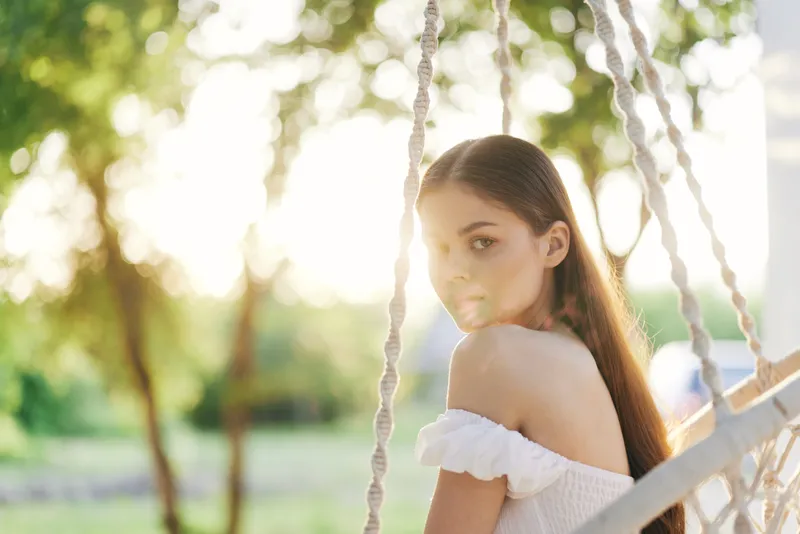 Young woman with long hair in white dress, sitting on a swing, surrounded by green trees, softly lit by sunlight, exuding serenity and calmness