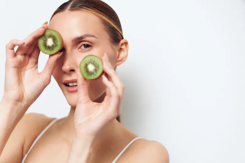 Woman applying kiwi skincare mask, placing fruit slices under eye, relaxed spa beauty self care concept, natural hydration treatment on clean background