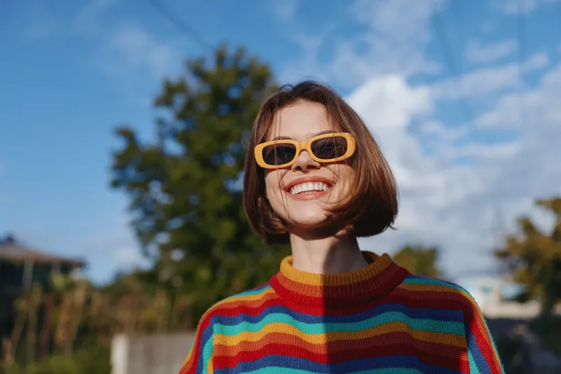 Young woman smiling outdoors on a sunny day wearing yellow sunglasses and a colorful striped sweater, short bob haircut, joyful travel lifestyle portrait in nature.