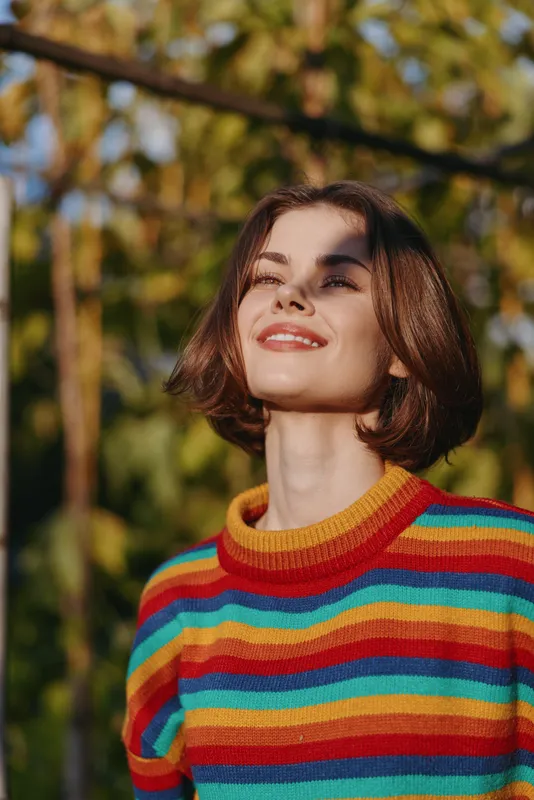 Young woman striped sweater smiling outdoors sunlight bob haircut casual outfit autumn joyful, standing in a sunlit garden looking up peacefully for lifestyle and fashion portrait.