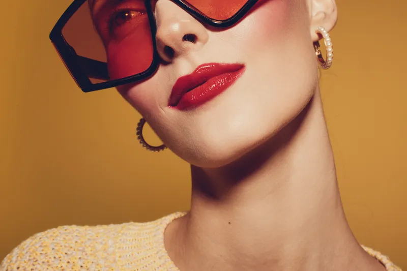 Close-up of stylish woman wearing large red sunglasses and gold hoop earrings with glossy red lipstick. Yellow background and knitted top create a modern fashion portrait concept.