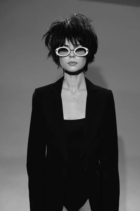 Stylish woman wearing white sunglasses and black suit with short hair in black and white portrait.
