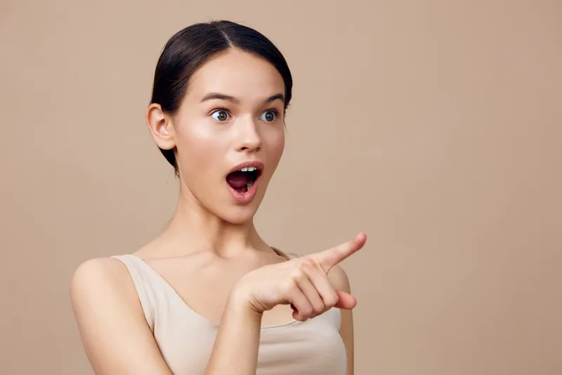 Surprised young woman with open mouth and pointing gesture against a neutral background showcasing excitement, curiosity, and surprise in a trendy outfit