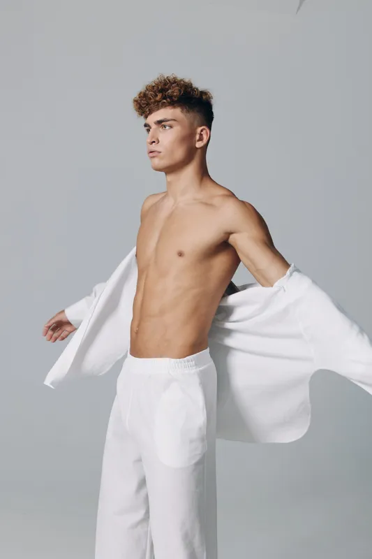 Young man with curly hair wearing white pants and open white shirt posing against gray background, showing toned body and confident expression in studio lighting.