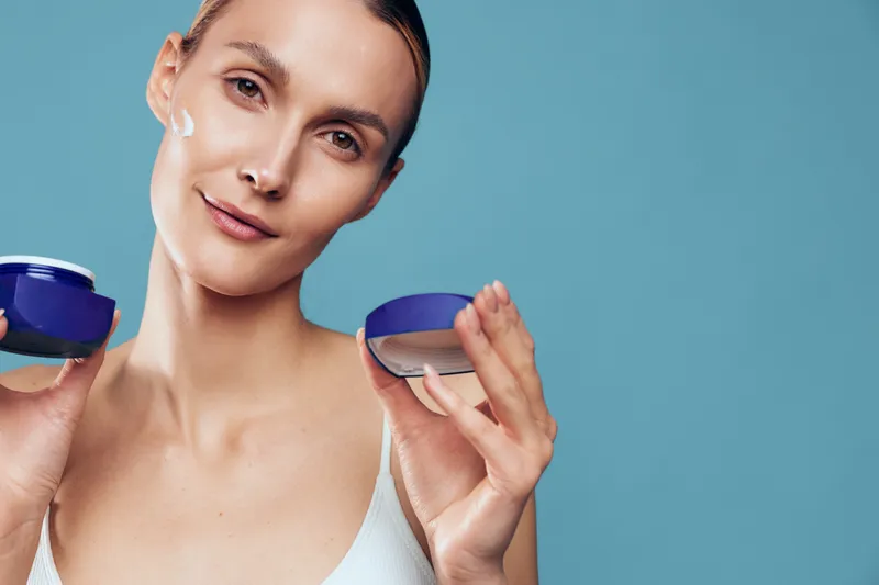 A young woman with clear skin applies cream on her face holding an open jar. The background is plain blue, emphasizing skincare and beauty routine concepts.