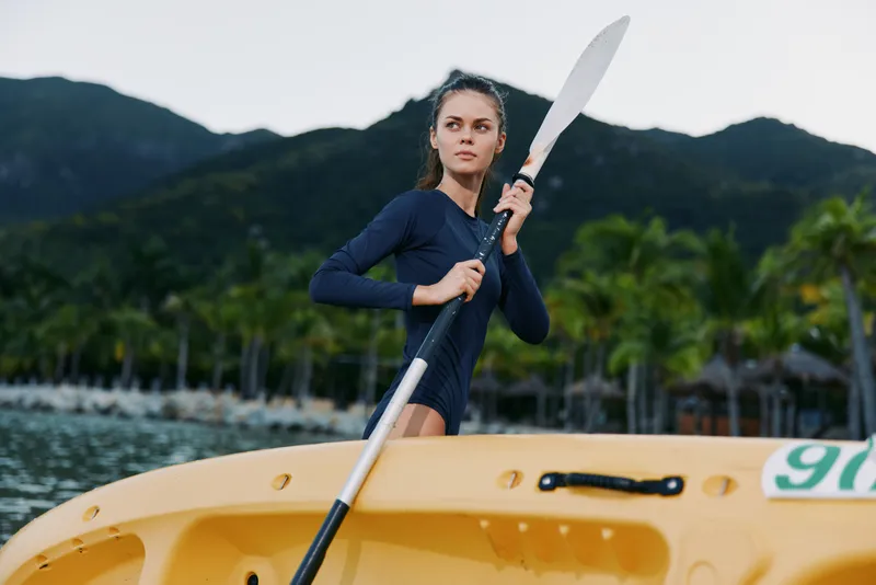 Young female kayaker stands beside a yellow kayak, gripping a paddle with focus. Scenic water, palm trees, and distant mountains create an adventurous outdoor sport portrait.
