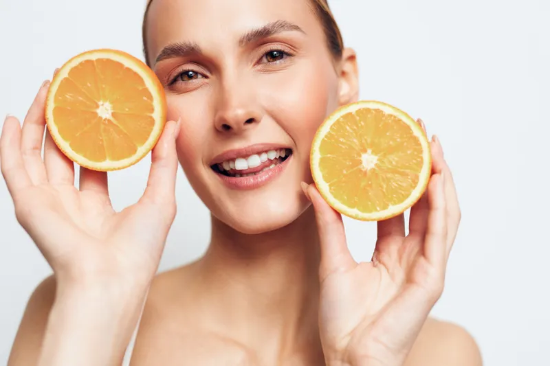 Young woman with clear skin holds two fresh orange halves near face smiles gently isolated on white background healthy lifestyle concept natural beauty and vitamin C source.