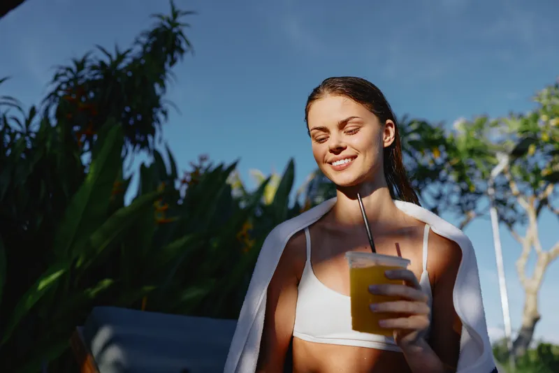 Young woman in a white bikini top smiles softly while sipping fresh orange juice outdoors in a bright tropical garden under a clear blue sky