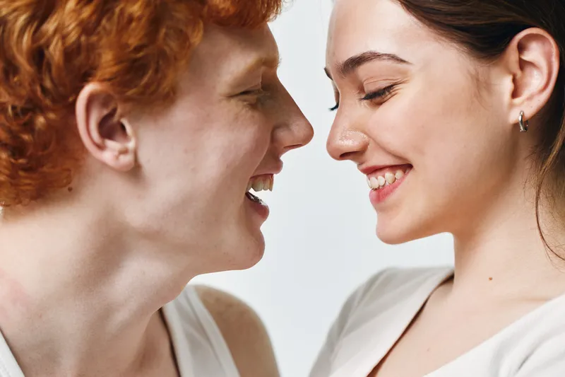 Happy young man and woman smiling and facing each other in closeup portrait with light background for friendship, youth and positive emotion indoors.