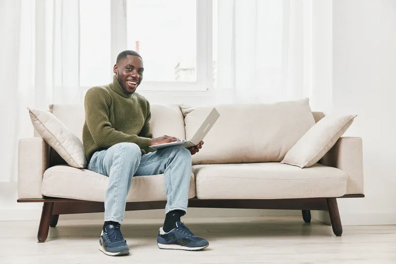 Happy Black man working on laptop from home, wearing casual clothes, seated on a light sofa with a bright interior, showcasing a modern lifestyle and remote work