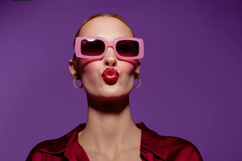 Young woman with pink sunglasses and red lipstick makes a kiss face on purple background. Fashion portrait with bold makeup and stylish accessories in studio lighting.