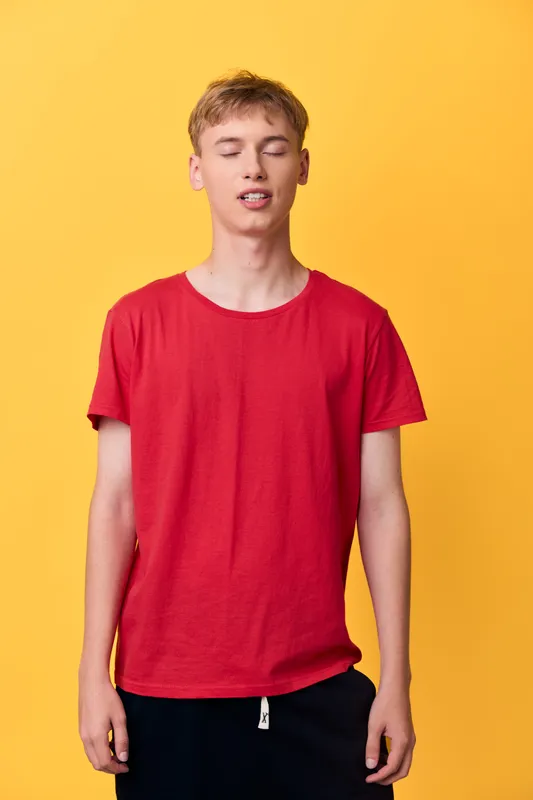 Red shirt youth in a casual studio setup against a bright yellow backdrop, relaxed pose with closed eyes, showcasing bold color, simple everyday fashion and contemporary style