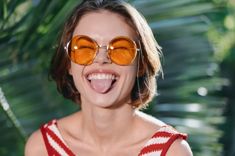A cheerful woman wearing round orange sunglasses smiles playfully in a sunlit tropical setting, creating a vibrant, carefree mood ideal for lifestyle and travel campaigns.