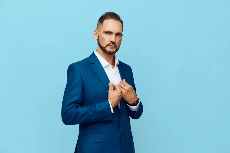 A confident professional man in a blue suit stands against a solid blue background, projecting focus and determination while adjusting his jacket with poise in an isolated color setting.
