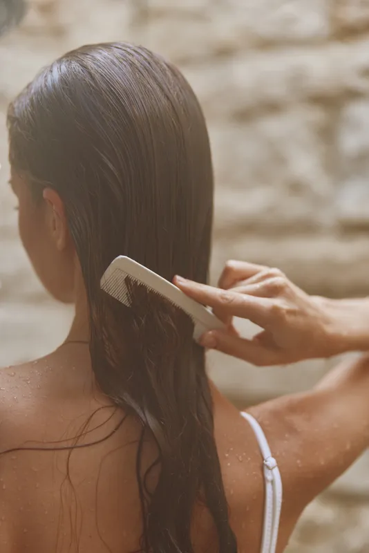Woman combing wet long hair gently with white comb against blurred natural stone wall background, featuring smooth texture and soft lighting.