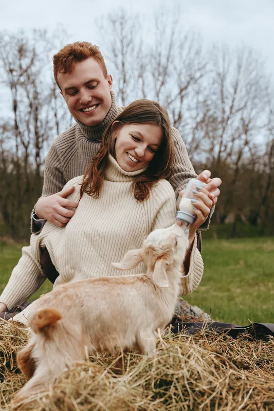 Joyful couple outdoors, smiling at a playful goat kid, soft sunlight, cozy sweaters, relaxed lifestyle, natural colors, grassy field backdrop, warm moment, gentle interaction, carefree atmosphere