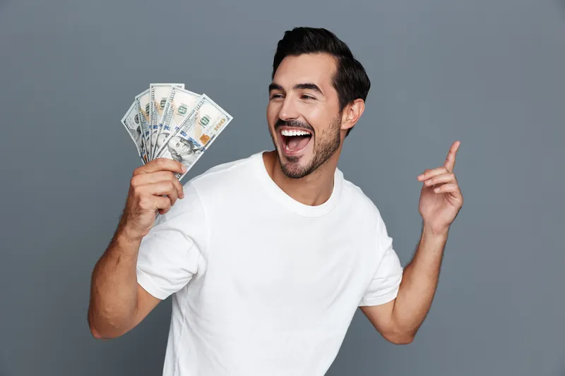 Happy smiling man holding cash in hand with excitement, showcasing his joy and celebration against a plain grey background. Opportunity and success concept.