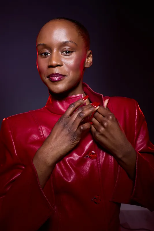 Confident black woman with dark skin wearing a shiny red leather jacket poses against dark background. Studio portrait of elegant African female model with short hair and bold makeup.