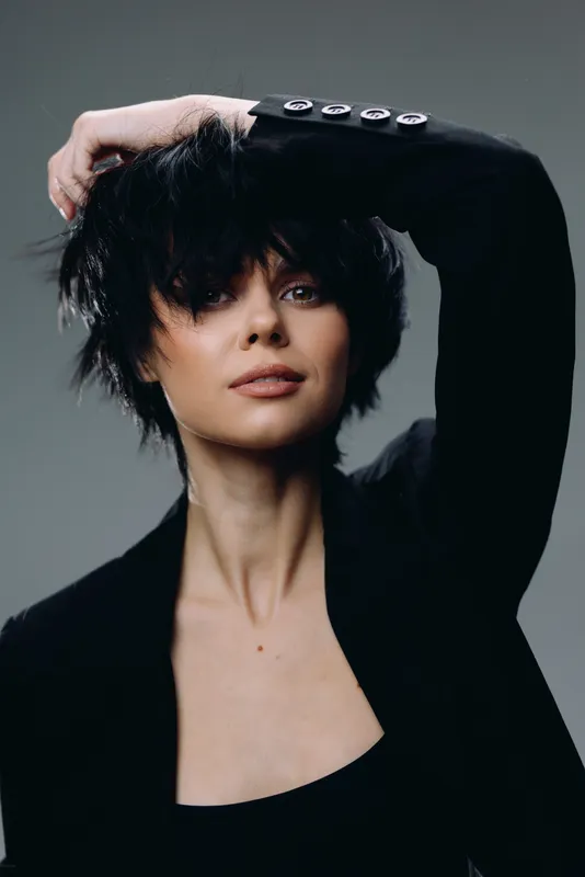 Stylish woman with short black hair wearing a black blazer posing with hand on head, neutral background, modern fashion portrait