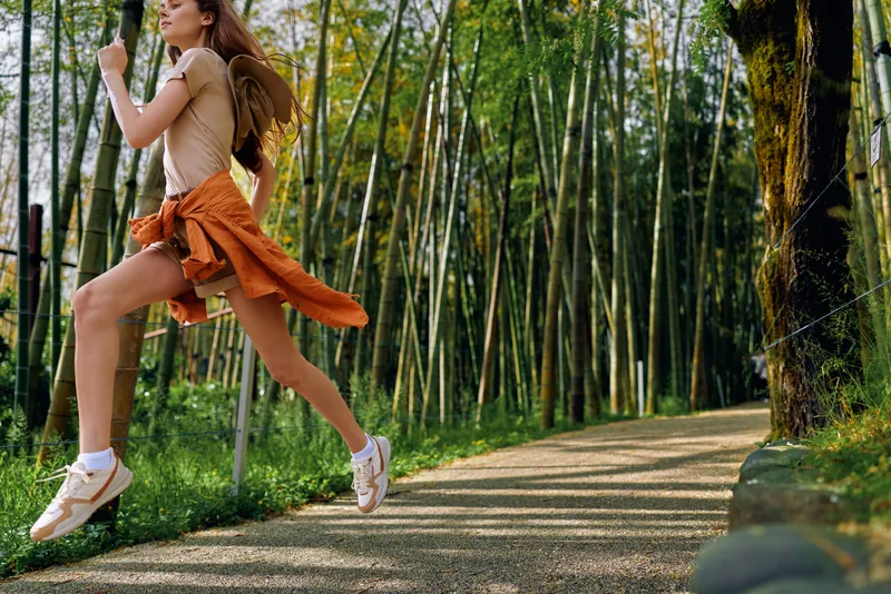 woman running and jump on a forest path, jogging with backpack and sneakers along a bamboo trail, active fitness motion in sunlight, outdoor exercise and healthy lifestyle in nature park