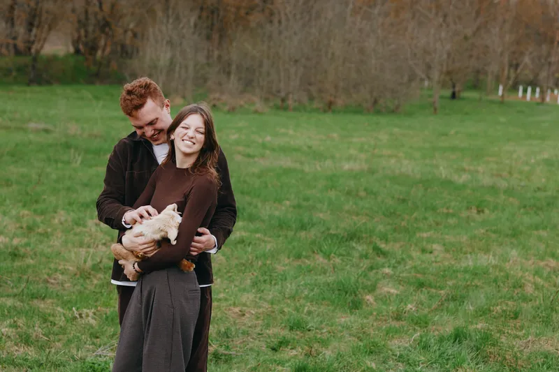 Young couple embraces outdoors as the woman holds a small goat in her arms on a green grassy field during a casual lifestyle moment.